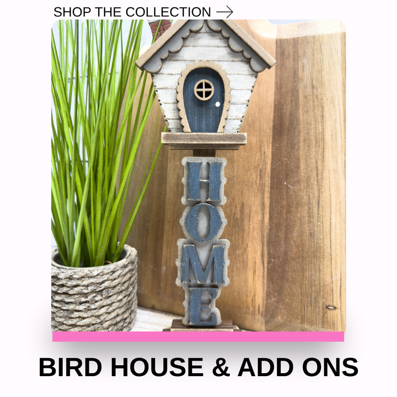 A decorative birdhouse-shaped sign with a vertical 'HOME' arrangement in blue-gray letters, shown next to a potted green plant and a wooden board. Text at the top reads 'SHOP THE COLLECTION ->' and text at the bottom reads 'BIRD HOUSE & ADD ONS'.