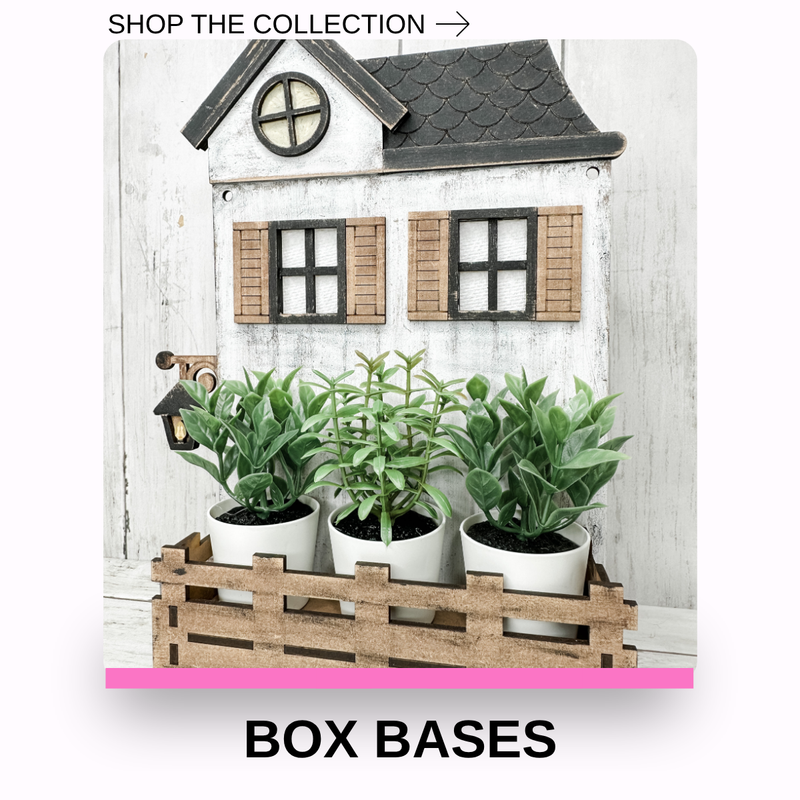 A decorative white house-shaped box base with a dark gray roof, brown shutters, and a wooden crate holding three potted green plants. Text at the top reads 'SHOP THE COLLECTION →' and text at the bottom reads 'BOX BASES'.