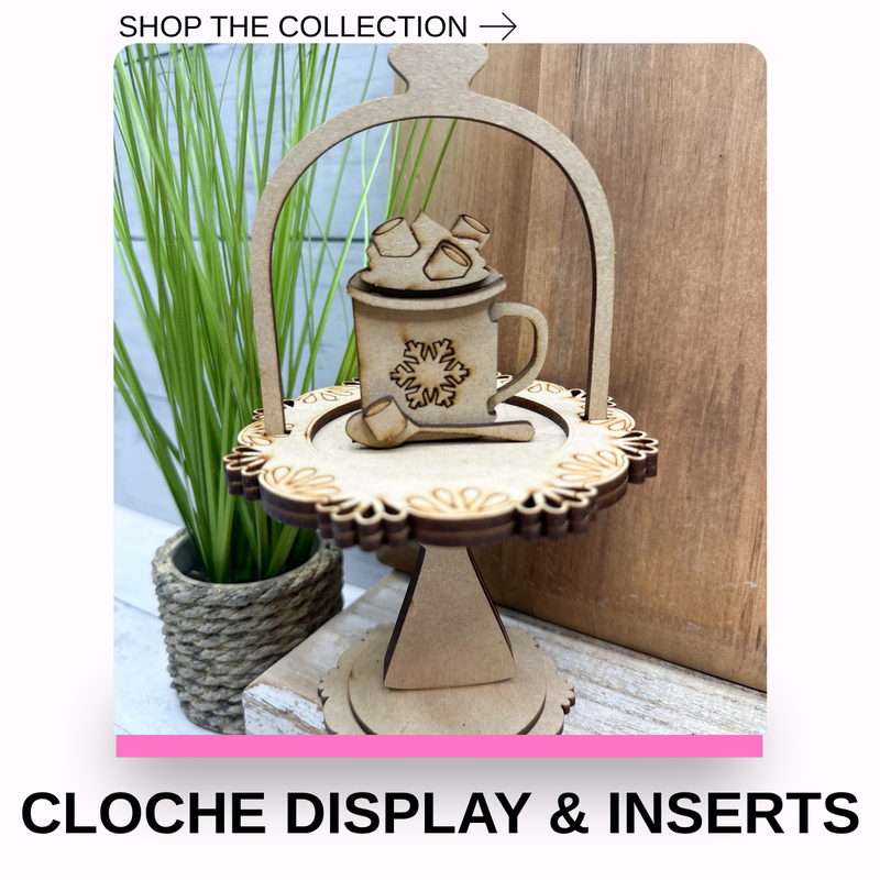 A wooden cloche display stand holding a small wooden mug with marshmallows and a spoon insert, shown in a styled indoor setting with a potted plant and wood panel background. Text at the top reads 'SHOP THE COLLECTION →' and text at the bottom reads 'CLOCHE DISPLAY & INSERTS'.