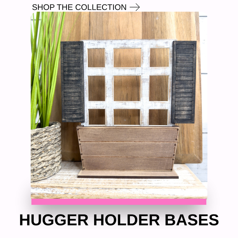 A wooden hugging holder base with a distressed white window design and dark shutters, shown on a tabletop with a potted plant and text overlays reading 'SHOP THE COLLECTION ->' and 'HUGGER HOLDER BASES'.
