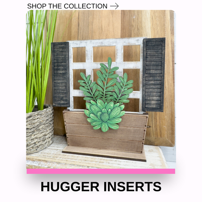 A decorative wooden window frame display with black shutters and a green succulent plant in a wooden planter, shown alongside tall green grass in a woven basket. Text at the top reads 'SHOP THE COLLECTION →' and text at the bottom reads 'HUGGER INSERTS'.