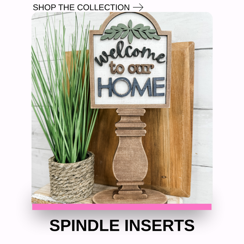 A decorative wooden sign reading 'Welcome to our HOME' mounted on a spindle-shaped wooden base, shown next to a potted green plant and wood panels. Text at the top says 'SHOP THE COLLECTION ->' and text at the bottom says 'SPINDLE INSERTS'.