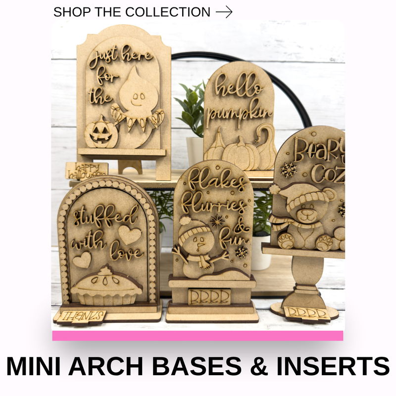 A set of laser-cut wooden arch stands with seasonal-themed engraved inserts, displayed on a white wood background with decorative plants. Text at the top reads 'SHOP THE COLLECTION ->' and large text at the bottom reads 'MINI ARCH BASES & INSERTS'.