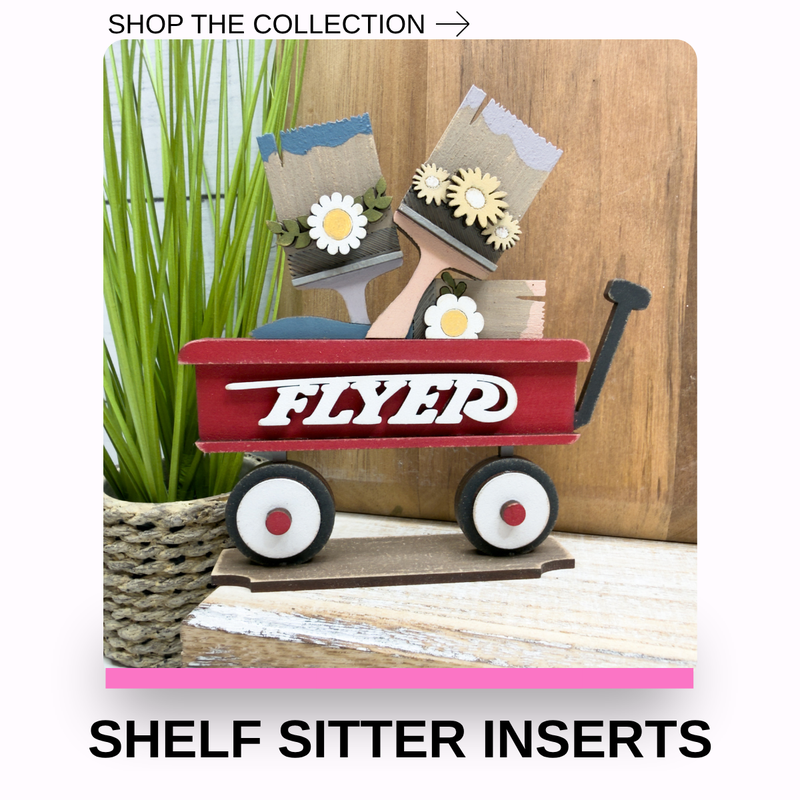 A decorative red wagon shelf insert with the word 'FLYER' in white script, holding small paint brushes with flowers inserts, displayed on a tabletop with a potted plant and wood panel background. Text at the top reads 'SHOP THE COLLECTION ->' and text at the bottom reads 'SHELF SITTER INSERTS'.