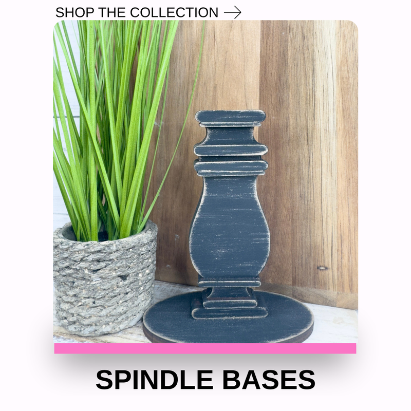 A dark navy blue distressed wooden spindle base standing on a matching round base, shown next to a potted green plant against a wood panel background, with text reading 'SHOP THE COLLECTION ->' and 'SPINDLE BASES' at the bottom.