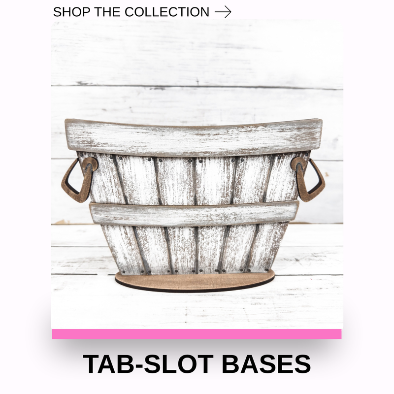 A rustic white-washed wooden barrel-style basket with distressed gray weathering and dark metal side handles, shown against a white wood plank background. Text at the top reads 'SHOP THE COLLECTION →' and text at the bottom reads 'TAB-SLOT BASES'.